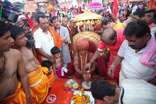 Outdoor ritual scene at a crowded Kumbh Mela-style ghat. A seated figure in a saffron-orange robe and deep orange turban sits on a gold throne with an ornate gold canopy overhead, wearing a long gold necklace and rudraksha-style beads. Before him a man in a red kurta with a tilak bends forward pouring liquid from a small brass vessel onto a white-cloth area, while a man in white shirt and glasses, with red tilak, watches. A brass thali holds yellow, red and orange powders and a marigold garland decorates a tray. Nearby, bare-chested priests in orange dhotis and devotees in white and orange crowd close, with photographers and a tall figure in white with rudraksha mala and orange shawl standing behind. Red flags and umbrellas fill the background.