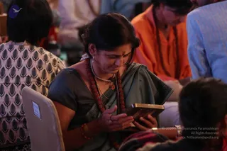 A woman in a grey-green sari with a dark border sits in a seated group and gazes down at a small gold-framed object she holds in both hands. She wears a red beaded rudraksha-style mala, a gold chain with a pendant, stud earrings and red bangles, with a bindi on her forehead. Behind her, a person in an orange garment with a beaded mala sits partly visible, and a seated figure in a white patterned top is in the left foreground. The setting appears to be a darshan session with participants seated closely together.