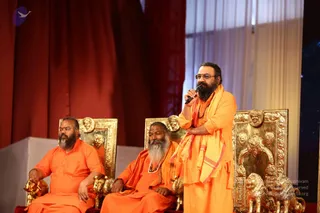 Three men in saffron-orange robes sit on ornate gold thrones with carved lion-head and lion-figure details against a red draped backdrop. The standing man on the right, with long dark hair and a full beard, wears rimless glasses, a saffron robe with a shawl, and holds a handheld microphone while speaking. The seated man on the left has a short beard and rests his hands on the throne arm. The seated man in the centre has a long white beard and wears a rudraksha mala.