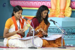 Two women sit cross-legged on a white floor singing devotional chant at microphones on stands. The woman on the left wears a white sari with a gold border, a yellow blouse and an orange shawl, with a black rudraksha-style mala and a small red bindi. The woman on the right wears an orange sari with a maroon blouse, gold bangles and beaded necklaces, with a white tilak on her forehead, and both hold printed sheets. Behind them a large painted deity murti's golden-clad feet rest on a turquoise and pink lotus pedestal.