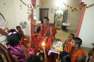 A photo of a seated figure in a red-orange robe and orange shawl on an orange-draped chair, holding a silver trident (trishul) upright in one hand. A red and white flower garland hangs around the neck, with gold bangles on the arms and rudraksha beads. Around the seated figure, several young boys in saffron-orange robes sit on the floor, and a girl in a pink floral dress and white hair ribbons watches while a woman in a white sari with gold border stands at left. A girl leans forward holding a lit oil lamp (aarti) above a silver plate of offerings with lemons, kumkum and vibhuti. Behind them, a shrine with framed pictures and flower garlands decorates the room, and a white-clothed table with fruit sits near a wall fan. The scene suggests a pada puja in a home, with the seated figure as Swamiji (probable) based on the trident, long hair, rudraksha and garland cues.