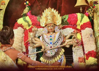A golden deity murti of Arunachaleshwara Paramashiva in a gilded crown, adorned with jewelled necklaces, earrings and a multicoloured gem-studded breastplate, stands framed by garlands of red roses, yellow and white jasmine flowers and green leaves. Multiple arms hold a trident and other ritual weapons. At the left edge, a woman in a white and gold sari with white body markings reaches toward the murti, offering worship. A golden bell hangs at the top right corner. The caption reads Arunachaleshwara Leela Alankaram Deeparadhana, Nithyanandotsavam 2021, Day 11.