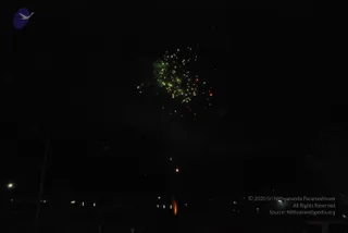 A night photograph of fireworks bursting in a dark sky over a distant, dimly lit area. A cluster of green and yellow-white sparks fans out from a central burst, with a trail of smaller glowing embers falling below. Faint street lamps and a utility pole sit at the lower left, and a small orange glow rises near the bottom centre. No people are visible in the frame.