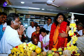 Photograph inside a clothing showroom with shelves of folded fabrics behind the group. A smiling man in saffron-orange robes with long dark hair, rudraksha malas and a thick red flower garland stands at centre-right, probable Swamiji, being welcomed. A man in white shirt and white dhoti on the left presses palms together in greeting. Nearby, a woman in a red sari and a woman in pink stand among a small crowd. In the foreground is a tray of bananas, apples, oranges and coconuts, and a tall silver aarti lamp with yellow flowers and a lit oil flame stands at right. Ceiling fan and fluorescent lights are overhead.