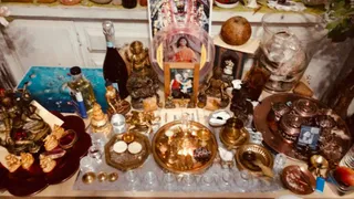 Overhead photo of a crowded home shrine table with many brass and bronze items. A brass puja thali holds small lamps and a brass kalasha-style vessel, surrounded by small glass cups and bowls. Several bronze deity figurines stand around the edges, including a seated figure and a ganesha-like form. A framed photo shows a robed figure in orange and red with a rudraksha-style necklace, placed near a glass bowl. A coconut sits on a shelf at the top right, and a blue cloth-covered panel lies on the left. No people are physically present in the scene.