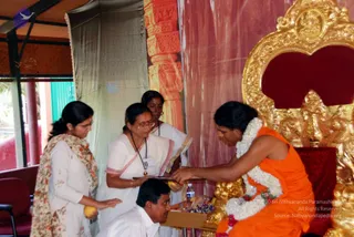 A seated figure in an orange saffron garment with a long white flower garland sits on an ornate gold throne with a red cushioned back, a gilded deity relief at the top. Three women in white saris and a man in white shirt stand and kneel before him, offering yellow fruit and a small plate of items on a wooden tray. The seated figure receives the offering with one hand extended. Behind is a red and cream draped backdrop and a stone-patterned pillar. A water bottle sits on the table. The white man kneels and handles the tray while a woman in a floral shawl holds a yellow fruit. Appearance matches the saffron and garland cues, so the seated figure is a person at an offering or darshan.