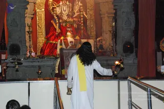 A photograph inside a temple shrine lit warmly from above. A person with long dark curly hair, seen from behind, wears a white dhoti-style garment with a yellow shawl draped over one shoulder and gold sandals; both hands hold a lit oil lamp or aarti flame toward a large deity murti. The murti is decorated with garlands and gold ornaments, set against red draped cloth and gold-framed niches, with brass oil lamps on the altar. Two or three onlookers are partially visible at the bottom edge. The visible figure's dress and long hair are appearance cues only; this is a puja or aarti scene in progress.