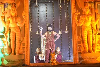 A photograph of a standing man in a stage-like shrine setting flanked by two large orange-lit guardian statues with multiple arms holding objects. He wears reddish-maroon draped cloth, a rudraksha mala and a large gold necklace, with a crown-like headpiece on his head. He holds a silver trident in his right hand and rests his left hand at his hip, standing before a golden throne-like seat with two carved lion figures and a striped cloth. Hanging bells and chains frame the dark backdrop.