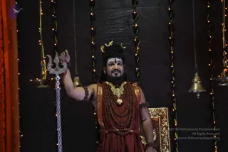 A man stands in front of a dark textured wall strung with small lights and hanging brass bells, holding a silver trident in his raised right hand. He wears a rust-red wrap, a heavy gold ornamental necklace over multiple rudraksha malas, and a crescent-moon headpiece in long dark hair, with white tripundra marks on his forehead. Oil lamps stand beside him and a gold-framed panel is visible at lower right. The image is a photograph from a darshan event.