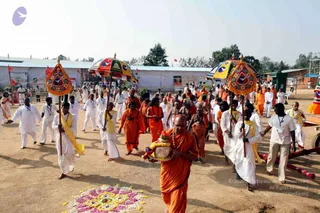 Outdoor procession on dry ground at a festival site with white tents and flags behind. Several people in saffron and orange robes and white dhotis walk in a line, with parasol-style umbrellas with blue and yellow canopies carried by men in white. The leading figure in front, wearing orange robes and a rudraksha mala, carries a brass kalasha or decorated offering vessel adorned with flowers. Behind him, white-clad volunteers with yellow shawls and lanyards walk alongside. A colourful flower rangoli design lies on the ground in the lower left. Ceremonial procession with saffron-robed participants and white-clad volunteers.