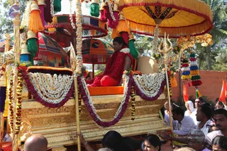 A procession on a gold-decorated ratha, a palanquin-style chariot, draped with white and purple floral garlands. A seated figure with long dark hair wears a red shawl and red garment and sits on an orange cushion beneath a large yellow canopy with fringe. Ornate poles, a trident-topped finial and multicoloured tassels frame the scene. Devotees in white and saffron stand along the sides, some with folded hands, and a white-clad woman is visible at right. Red bougainvillea-style flowers and orange flags appear in the background beside an orange-brown wall.