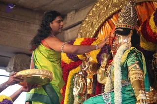 A photograph of a temple ritual at a deity murti. A man with long, swept-back dark hair and a bare upper body wears a lime-green silk dhoti with a red-and-gold border and a pearl necklace and bangles. He reaches toward a gold-faced murti dressed in a green garment with gold armour, a jewelled crown and a white flower garland. Behind the murti are dense bands of red, yellow and purple flowers and gold-embossed fabric. On the left, another person holds a gold plate.