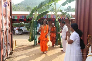 A photograph of a gateway decorated with banana trunks and leaves, flanked by red shipping containers. A man with long dark hair, wearing an orange dhoti and a large white and orange floral garland, smiles and raises a hand in greeting as he walks barefoot through the gate. Behind him a shaven-headed man in orange robes with a rudraksha mala follows. On the right, a woman in white with a black bag stands with palms pressed together in namaste, and a man in white shirt and white dhoti stands nearby. Strings of coloured bunting hang along a building behind, with a silver car and wire fencing in the background.