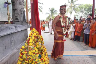 Outdoor temple courtyard with palm trees and a grey stone shrine wall on the left, a brass oil lamp on a stand and a red cloth hanging. A smiling man in a rust-red dhoti and shawl, wearing a red and white flower garland, a red headband and gold armbands, stands beside a large mound of yellow and red flower garlands that covers a figure. Behind him a white-clad priest and a line of saffron-robed boys and young men in orange garments and rudraksha malas stand watching, several with hands folded. The man appears to be a person, given the garland and ornate dress, though his face is not used for identification.
