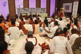 A large seated satsang in a carpeted hall with lavender patterned walls. Many devotees sit on the floor in white clothes with orange-brown shawls, some wearing rudraksha malas. In the foreground a woman in a white embroidered top and a woman with dark hair sit among them. On a raised red and gold throne at the far right, a figure in saffron-orange robes with a crown sits under a golden parasol, beside a trident and a brass lamp. Behind the seated crowd a green-draped table holds framed photographs arranged on boards, and several monks in orange robes sit near it. Count of visible people is large, roughly 60 to 80.