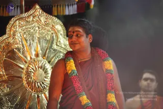 A photograph of a man in a maroon-red robe standing before an ornate gold-toned metal backdrop with a radiating sunburst pattern. He wears a multicolour garland of pink, yellow and teal flowers over his shoulders, a red bindi and white-and-red tilak on his forehead, and a rudraksha-style mala at his neck. Smoke or incense haze drifts behind him, and a second, blurred man in an orange-red cloth is partially visible at the right edge.