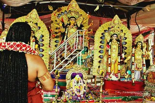 A photograph of a shrine during Navaratri at an ashram temple. In the foreground, a figure seen from behind with long dreadlocked black hair, wearing a red-and-white patterned shawl and a red garment, with a gold armlet on the upper arm, faces the altar. Behind them stand large, ornate golden deity murti with multiple arms, holding weapons, with gold halos edged in blue eyes, and a lion mount visible at the base. Flanking statues of goddesses wear yellow and gold garments and garlands. Marigold and flower offerings lie before a small shrine, with brass oil lamps and rose petals on the floor. Pink and white draped fabric forms the tent ceiling.