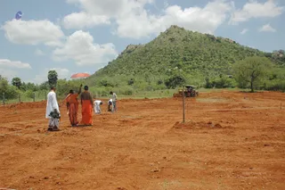Open reddish-brown earth at a construction site beneath a blue sky with cumulus clouds. A rounded green hill rises in the background with scattered trees and palms. A tractor works at right near a fence line. Four people in the foreground walk and stand on the cleared ground: a man in white dhoti and shirt holding a small object, two figures in orange-saffron cloth with one raising an arm, and a bare-chested figure in orange dhoti. Two more people in white bend over, measuring or surveying along a taut white string stretched on wooden stakes. A red umbrella stands at left. The a person candidate is not confidently identifiable from appearance, so the role is given as a figure in saffron cloth.