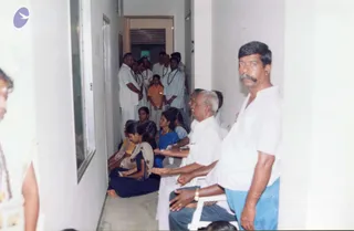 A photograph of a white-walled corridor at a healing centre, with people waiting in line. On the right, a man with a thick grey moustache in a white shirt and blue lungi stands beside seated devotees with palms extended. Seated in white plastic chairs are an older man in a white shirt and a woman in a blue sari, and a younger woman in a cream top sits cross-legged on the floor. Behind them, a group of men in white shirts and dhotis, some wearing rudraksha malas, stand in a line near an open doorway, with a small boy in an orange shirt among them. Several people are visible in the background, and a partially visible figure with a garland stands at the left edge. No a person candidate can be clearly identified by appearance.