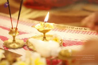 Close-up photo of brass ritual items on a pink and white woven cloth. A brass oil lamp with a lit flame sits on a brass tray, flanked by two brass incense holders with burning incense sticks. White and yellow frangipani flowers lie in the foreground and on the tray. A blurred hand and red fabric appear at the edges. No people are clearly visible.