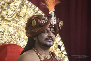 A man seated before a large gold ornate backdrop speaks into a microphone at right. He wears a rust-brown turban decorated with gold and jewelled ornaments, a sprig of orange feathers, and a dark red-brown wrap over a bare chest. Layered gold and bead necklaces hang at his neck, and a red cushion appears at lower left.