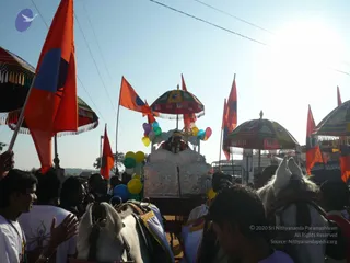 A festival procession on a sunlit road during the Jayanti Brahmotsavam celebrations. A decorated chariot with a white and silver ornamented body and a small umbrella canopy is drawn by white horses with gold-trimmed harness. Balloons in yellow, green, blue and pink are tied to the chariot, and a figure sits under the canopy. Orange and red flags on poles and several multicoloured ceremonial umbrellas with tasselled fringes accompany it. Devotees in white shirts and dhotis walk alongside and crowd the road, several raising hands in greeting. Blue sky and a distant tree line form the background.