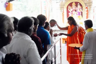 A temple interior where a long-haired figure in orange sannyasi robes stands before an ornate shrine with deity murtis draped in red and decorated with gold. The robed figure is placing a hand on the head of a devotee in white shirt and pants, who holds his palms together in namaste. A line of devotees in white, blue and maroon shirts waits behind a metal railing. On the right, an attendant in white with a yellow shawl holds an orange tray. The figure shows distinctive long hair and orange robes; Swamiji (probable) performing blessing at the shrine.