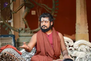 A bearded man with long dark hair and white forehead markings, wearing a rust-brown shawl over a maroon top, sits on an ornate silver chair with elephant carvings and gestures with one open hand. Behind him a bronze dancing Shiva figure stands against a deep red wall, and a pillar is visible at right. A leopard-print cloth lies across a lap at lower left. Other people are only partly visible at the bottom edge.