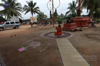 Outdoor photo of a temple courtyard with reddish-brown earth and coconut palms. A single seated figure in orange robes with red and white beaded necklaces sits cross-legged on a mat in front of a circular floral-lined ring and small oil lamp, a lotus and floral kolam drawn on the ground. Along the path, rows of stone deity murtis and grey sculptures stand on red-bordered bases, with a tall ringed pole with red and orange banners and a large orange-red stone shrine structure at right. A silver SUV is parked at left and a poster of deities hangs on a wall behind. Two pigeons stand on the ground.