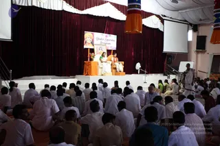 A seated audience of many devotees, mostly men in white clothing, faces a raised stage with maroon velvet curtains and orange-and-white drapery. A banner reading "Bhakti Spurana Program" hangs above two women seated in white armchairs on an orange-draped platform; one wears a white sari with green trim and the other a white sari with an orange shawl. A framed portrait stands beside them with a vase of flowers. Orange and multicolour hanging banners, projection screens, a fan and a standing camera on a tripod complete the hall. A man in white on the right stands near the stage.