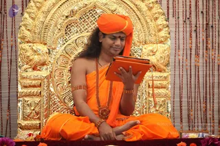 A seated man in saffron-orange robes and matching turban sits cross-legged in front of an ornate gold carved throne-backdrop with lion-like motifs. He wears a long rudraksha-style bead mala, gold-toned bangles and an armlet, and a red bindi on his forehead. He holds an orange tablet device in both hands and looks down at it, reading. A beaded curtain hangs to the right, with flower garlands and pink and purple flowers at the bottom edge.