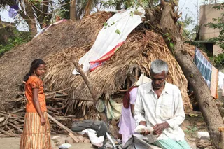A photograph of a damaged village scene after a cyclone. A thatched hut with a torn white tarpaulin and a faded red banner strip stands behind a stacked pile of wooden sticks. A young girl in an orange blouse and striped skirt stands at left looking toward the camera. An older man with grey hair, wearing a white checked shirt and green striped lungi, sits on a motorbike counting paper bills. A woman in a pink garment stands partly hidden behind him, and laundry hangs on a line at right.