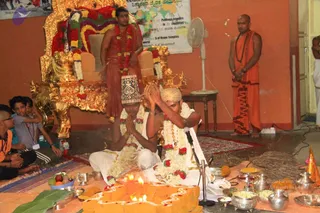 A photograph of a homa or puja ceremony in a temple hall. Two men in white dhotis and white shawls sit cross-legged on the floor before an orange-coloured ritual fire pit lit with oil lamps, their palms pressed together in prayer; one wears a decorated cap and both wear white flower garlands with red blooms. Behind them stands a man in a reddish-brown dhoti with a red garland, beside a gold-and-red ornate throne draped in flowers. On the right a man in saffron-orange robes and a rudraksha mala stands with hands folded. Brass vessels, fruit, a banana leaf and a microphone are on the floor. Several young boys sit at the left edge. A world map and a poster on the orange wall are behind them.