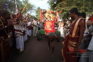 A street procession in a tree-lined road during a Kumbh Mela-style religious event. In the foreground, a bare-chested elderly man with grey hair prostrates on the road, hands forward, beside a red flag bearing a portrait of a saffron-turbaned figure. Robed devotees in white and red-orange dhotis with gold borders stand on both sides, one raising a yellow staff and clapping. Behind, a green tractor-style cart draped with red flags carries a decorated golden canopy and garlanded deity murti, with a uniformed police officer walking alongside. Several onlookers with cameras watch from the roadside.