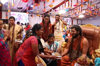 A festive hall decorated with hanging colourful paper lamps and diyas under a white canopy. A bearded man in orange robes and a leopard-print shawl with long rudraksha malas sits on a gold-trimmed brown leather throne at right, smiling, with a golden lion-head armrest visible. Beside him a woman in a red and orange patterned sari and black shawl bends forward offering a tray with lamps and gold items, while a young girl in a white and blue sari and red beaded necklace leans in to receive it. Behind them a shaved-headed woman in an orange-brown garment with a long rudraksha mala stands, and a man in a white dhoti with a brown shawl and rudraksha mala looks on. At left, women in gold and green saris and a woman in pink stand with hands folded, and a large video screen shows figures in the background.