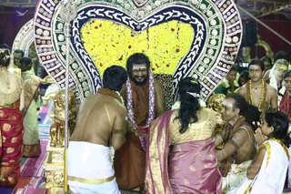 Photograph of a ceremony in a decorated hall. A bearded man with long dark hair sits smiling on a throne backed by a large heart-shaped frame of yellow marigold garlands and red-and-green patterned borders, with a leopard-print cloth on the seat. He wears a rust-orange robe and a multi-coloured flower garland. In front, a bare-chested young man in white dhoti with sandalwood markings bows toward him, and a woman in a pink and gold sari kneels nearby. To the right, a man with ash markings and a rudraksha necklace, and a young boy in orange with a beaded garland, sit in attendance; a girl in white with gold border folds her hands. A golden trident stands at left. Several other seated people in saris and white clothing are visible at the edges. Ceremonial puja setting.