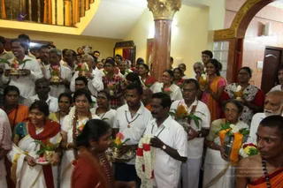 Photograph of a crowded indoor hall with yellow walls, a tall ornate brown pillar and a wooden staircase railing. Many devotees in white clothes stand holding brass pots and plates with mango leaves, pink flowers and fruit, and white and red garlands. In the foreground a woman in a red-brown sari and a man in a white shirt with a garland face the viewer. Several women wear white saris with orange borders and a young woman in orange-and-white dress stands at right. A man on the far right wears saffron robes and a rudraksha mala, with a tilak on his forehead. No clearly identifiable a person is visible.