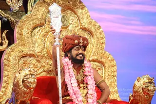 A smiling bearded man in a rust-orange turban with a jewelled ornament and a rust-orange robe sits on an ornate gold throne carved with floral scrollwork, flanked by gold lion heads with red upholstered seat. He holds a tall silver ceremonial staff in his raised right hand and wears a long garland of pink lotus flowers along with heavy gold necklaces and gold bangles. The background is a soft pink and violet backdrop. Watermarked copyright text appears in the lower right corner.