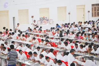 A large hall with white walls and tall cream-colored doors where many devotees sit at long white folding tables on red plastic chairs, eating from plates. Most wear white clothing, and several women wear saris in pink, orange and green. A standing woman in white on the left watches over the seating, and a framed picture of a seated figure with garlands, flanked by two smaller framed photos and small lamps, is placed on a table at the back. The scene looks like a communal meal in an ashram setting.