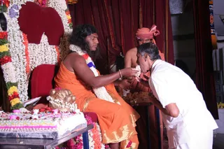 A seated figure in an orange saffron garment with gold trim, wearing a long white and red flower garland and a blue-and-white garland, sits on a red throne-like chair decorated with white jasmine and red and yellow flowers. He has long dark swept-back hair and a dark beard. Beside him a man in an orange turban and orange robe stands behind a table of offerings. In front, a man in white shirt and white trousers bends forward to receive a sweet from the seated figure's hand. A large decorated cake with pink and white icing and red berries sits on a table at left, and red drapes hang behind.