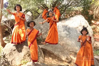 Six young girls in orange saris with dark red borders pose on and between large grey granite boulders outdoors, with green trees and foliage behind them. Several girls wear rudraksha-style beaded malas and necklaces; one wears a gold-toned headband. Some lift their arms or stand with hands raised in a playful pose on the rocks. No a person candidate is visible in the scene.