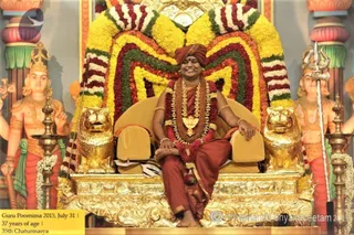 A photograph of a seated man on an ornate gilded throne decorated with a mass of yellow, red and white marigold garlands arranged in a fan behind him. He wears a rust-red turban, a rust-red robe, a dense rudraksha mala with a large gold-toned pendant, and a red flower garland, and smiles with hands resting on the armrests. Flanking him are golden lion figures on the throne base, two painted standing deity figures holding a trident, and brass oil lamps in front. A caption in the lower left reads Guru Poornima 2015, July 31, 37 years of age, 35th Chaturmasya.