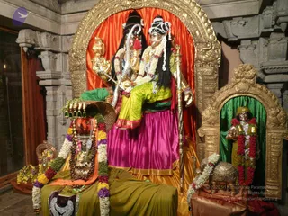 A decorated temple shrine with a golden arched niche framing two large deity murtis seated together on a throne. The central pair wears magenta and lime-green silk draped garments, white and yellow flower garlands, and a silver crown-like headpiece, one holding a bow-like staff. Beside them stands a smaller standing murti in a green-curtained niche wearing a green robe and red garlands. A brass kalasha or lamp stand, marigold garlands and a green cloth decorate the foreground altar. The shrine is carved stone with gold trim.
