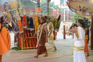 A photograph of a temple procession at an outdoor ashram courtyard. In the centre, a bearded man in a rust-brown dhoti and red-and-gold turban, with a long garland of white and pink flowers and gold ornaments, walks barefoot beside a companion in a white dhoti and sleeveless top with gold borders. Behind them, a cameraman in white dhoti with gold border films, and a figure in orange robes stands at left. A large ornate umbrella with gold finials and tasselled fringe is held overhead at right, and a rack of colourful ceremonial flags with silver tassels stands behind. a person candidate in a turban with a garland and gold necklaces walks in the procession.