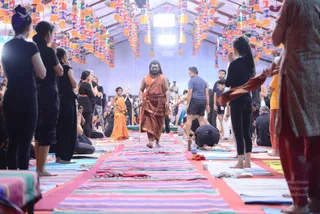 A large tent hall decorated with hanging colourful paper lanterns and tassels. A central man with long dark hair, a full beard and an orange-saffron wrap walks barefoot down a runway of striped woven mats and rugs, with a cord or wrap around his waist. Around him, many people in black clothing stand with palms pressed together in anjali, while others sit or bow on mats; a child in an orange sari stands near the centre. A man in a grey t-shirt and dark shorts stands to the right, and a woman in a dark outfit holds up a saffron cloth. The scene suggests a satsang or yoga gathering, with a visible bottle and cups on the floor.