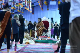 A photograph of a session in a large tent hung with tassel decorations. A man with long dark hair and a beard, wearing an orange-brown robe, sits on an ornate silver-and-red throne holding a microphone and smiling. Around him, participants in black athletic clothing stand with hands raised to their faces on striped rainbow mats, while a man in white robes and a seated attendant sit on the floor nearby. A red water bottle lies on the mat in front, and a standing figure in purple is visible in the background. The scene reflects a satsang or yoga session, and the appearance-based label for the seated man is probable Swamiji (throne, long hair, orange-brown robe, microphone).