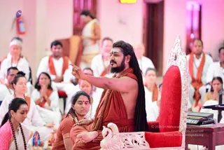 A seated man in saffron-red robes with long dark hair, a full beard, a rudraksha mala and a forehead tilak sits on an ornate white-and-red throne with carved swan arms, pointing with his right hand toward the audience. A woman in a saffron sari sits beside him on the floor. Behind them, rows of devotees in white clothes and saffron shawls sit attentively, some with hands folded. A keyboard and microphone sit on a side table at right. The room has pink-lit walls and a wooden door. a person: the seated teacher on the throne has long swept-back hair, saffron robes, a mala and a ceremonial throne, consistent with a discourse setting.