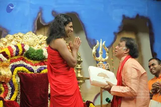 Photo on a stage with a blue painted backdrop. A long-haired man in a red draped robe stands with palms pressed together in namaste, smiling, beside an ornate gold-and-red floral throne decorated with marigolds, roses and green leaves. Beside him, a man in a peach kurta with a red shawl offers a white gift box with a gold tree motif, while a golden trishul and brass lamp stand nearby. Another man in orange clothing smiles at the right edge. The long-haired figure, in a red robe with a gold-beaded necklace and a Swamiji (probable) appearance, is greeted in a ceremonial exchange.