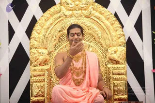 A seated man in pink-saffron ceremonial robes sits on an ornate gold throne with a carved arched back and lion-like figures at its base. He has a red bindi on his forehead, multiple tiers of gold beaded necklaces and gold armbands, and his eyes are closed while he raises one hand to his chin. The backdrop is a black and white diagonal lattice pattern.