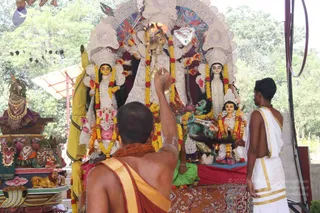 A photograph of an outdoor shrine dominated by a large white and red deity murti with multiple goddess figures adorned with yellow and red marigold garlands, a pink Ganesha figure at the front, and a green seated figure. A priest with a shaved head, wearing an orange-red shawl, raises a brass aarti lamp with a flame toward the deities. To the right, a man in white dhoti and shawl with gold borders stands with hands near his waist. Lions and a serpent-like sculpture decorate a side platform, and trees surround the open-air setting.